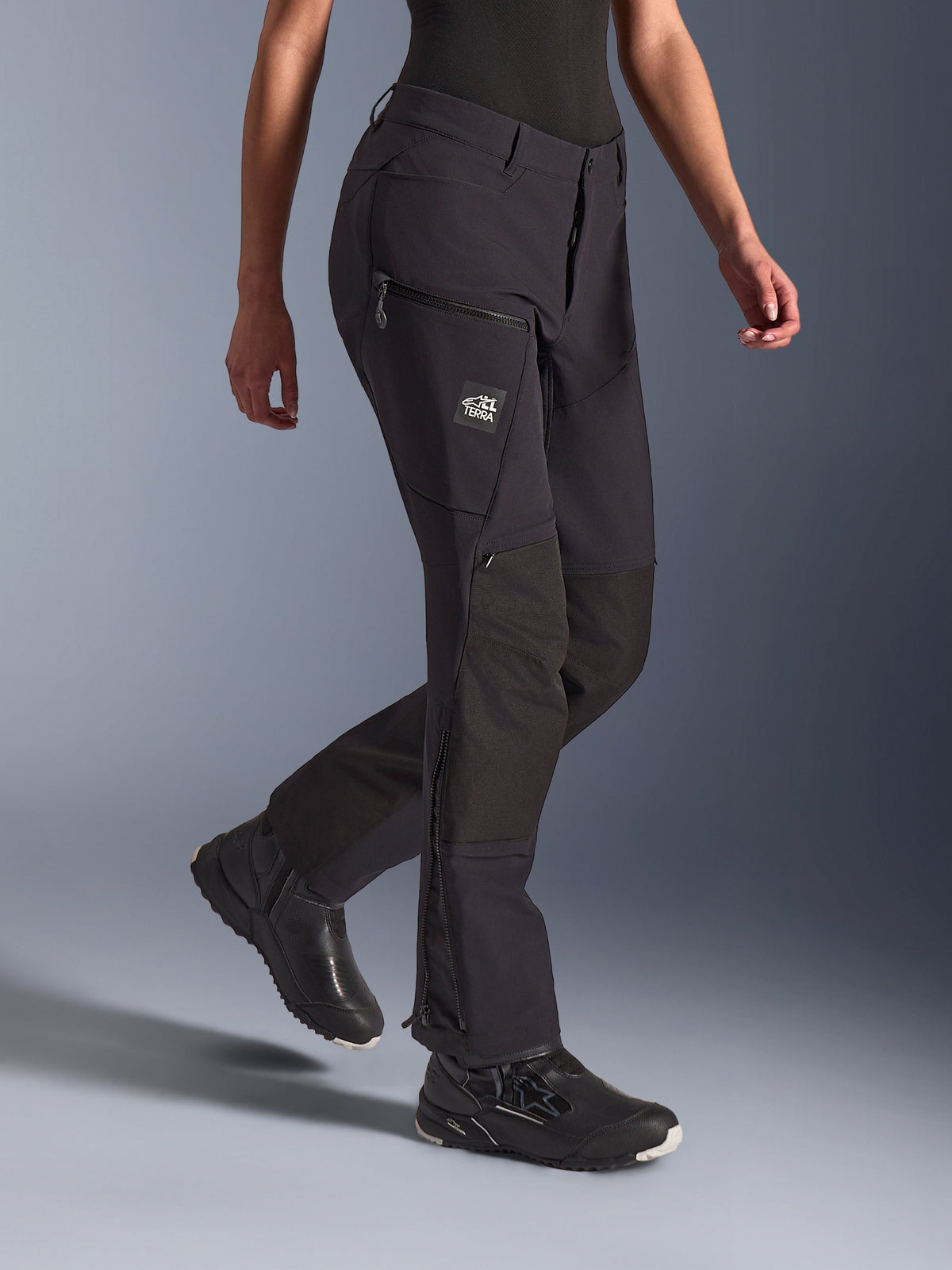 Pantalones Alpinestars Stella Flex-AST Explorer para mujer, negro, pantalones de motociclismo adventure con construcción de textil elástico y refuerzos de ripstop en las rodillas, bolsillos cargo con cremallera y marca TERRA, y cremalleras en la parte inferior de la pierna, usados con calzado de moto negro.