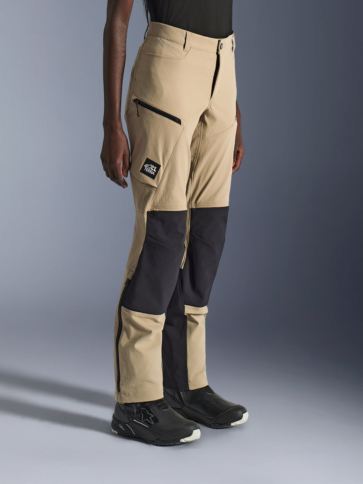 Mujer con pantalones Alpinestars Stella Flex-AST Explorer, pantalones de moto adventure en color arena y negro con paneles de ripstop negro en las rodillas, bolsillos de carga con cremallera, parche con el logo 'ALL TERRA' y cremalleras en la parte inferior de la pierna.