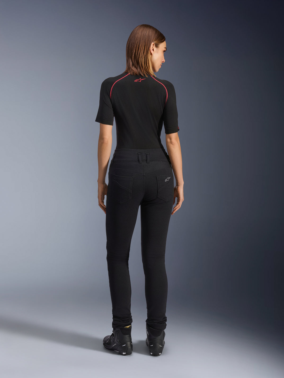 Leggings Banshee para mujer de Alpinestars, leggings de motociclismo negros, vista trasera que muestra el ajuste ceñido y el tejido elástico anatómico, con bolsillos traseros y detalle del logotipo, usados con una capa base negra y calzado de conducción, equipamiento de moto urbano para mujer