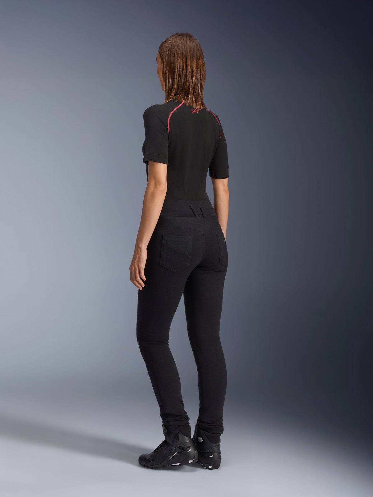 Leggings Banshee para mujer de Alpinestars, negros, vista trasera que muestra el ajuste anatómico elástico ceñido y los bolsillos traseros, combinados con un top negro de manga corta y botas de moto, equipamiento de moto urbano y elegante para mujer