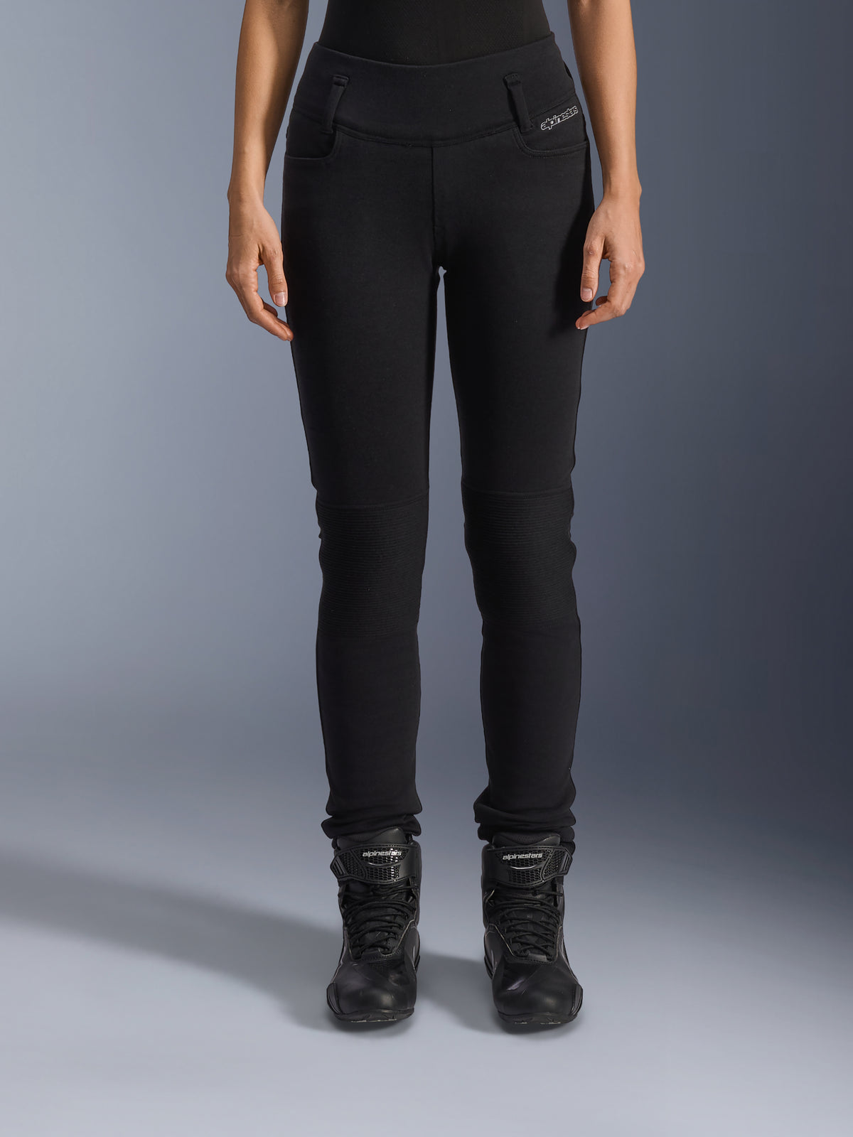 Alpinestars Womens Banshee Leggings, pantalones de moto negros, diseño ajustado con paneles de acordeón en las rodillas, combinados con botas de motociclismo negras de caña alta, equipamiento urbano con un estilo casual