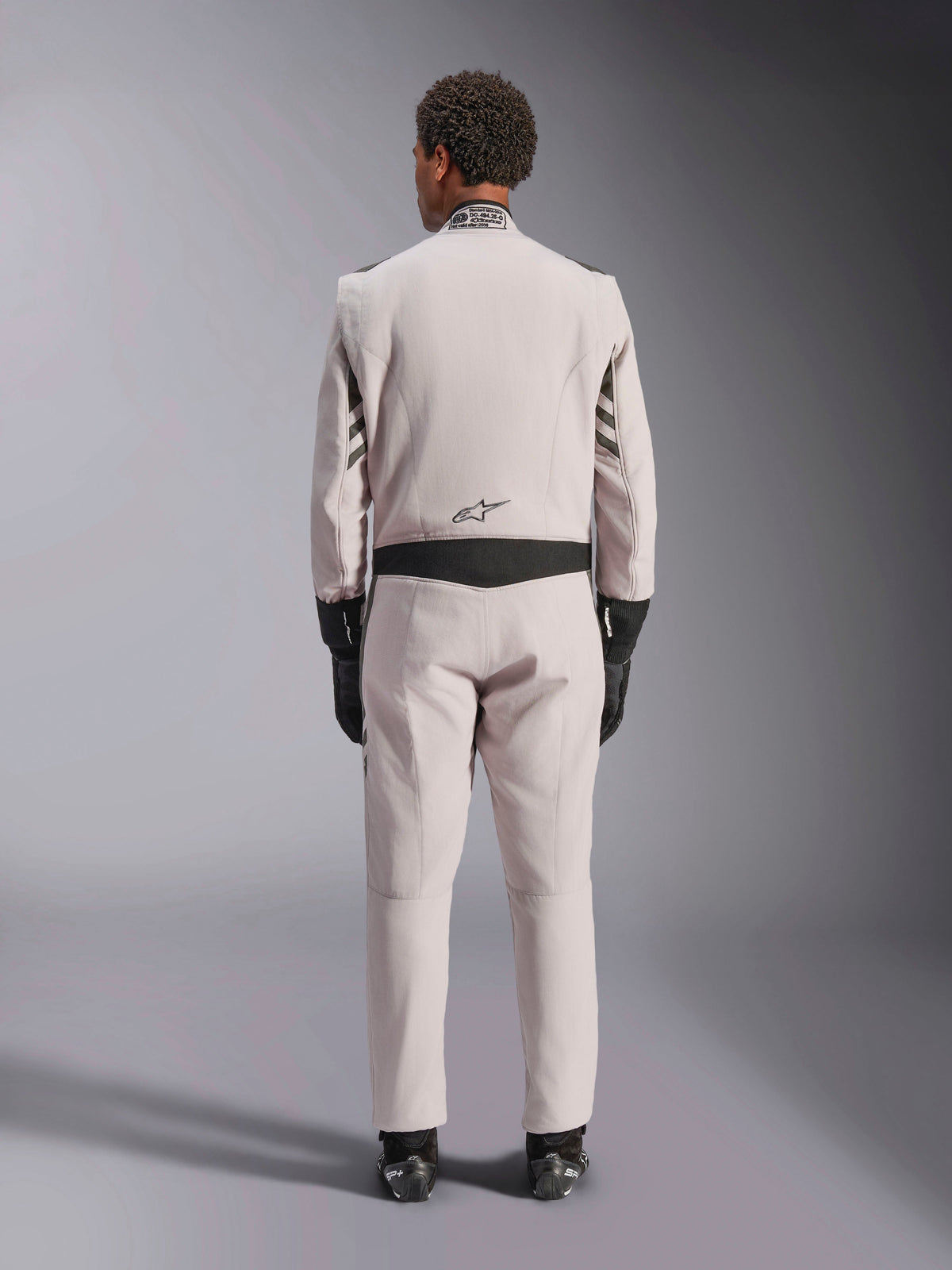 Alpinestars GP Race V3 Suit FIA, Silver Anthracite, vista posterior de un traje de automovilismo de una pieza usado con guantes y botas negros, con paneles elásticos negros en la cintura y las mangas, diseño profesional de automovilismo