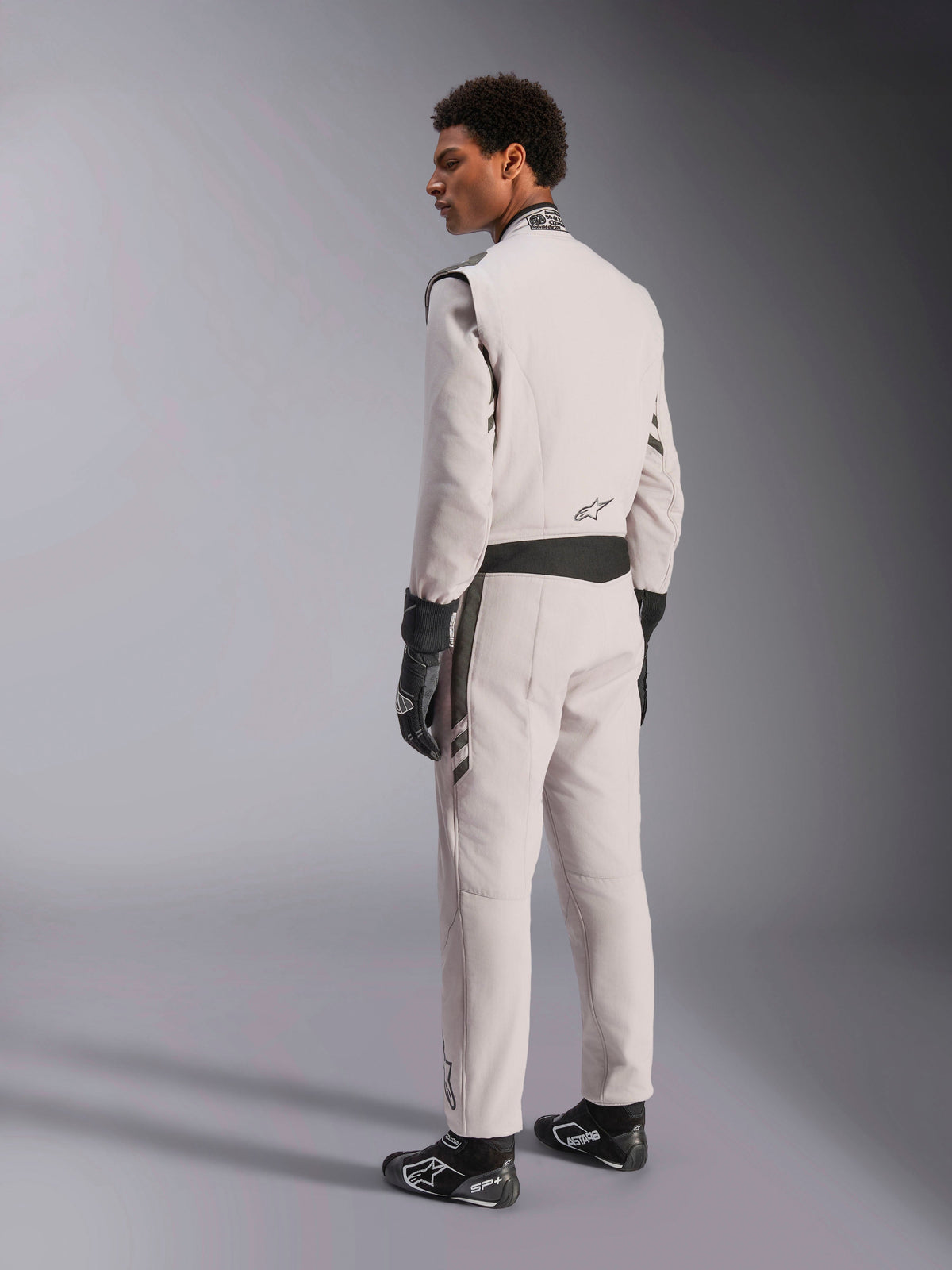 Traje Alpinestars GP Race V3 FIA, mono de carreras en color Silver Anthracite, vista lateral y trasera, usado con guantes y botas de carreras negros, con detalles en gris oscuro en la cintura y los hombros, equipamiento protector profesional para automovilismo.