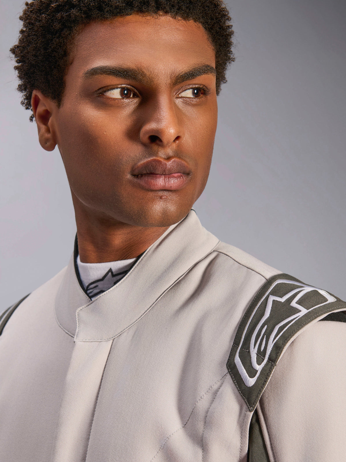 Modelo vistiendo el traje Alpinestars GP Race V3 FIA en Silver Anthracite, toma de primer plano centrada en el torso superior y el hombro, destacando la hombrera de extracción y el cuello ergonómico, equipo profesional de automovilismo
