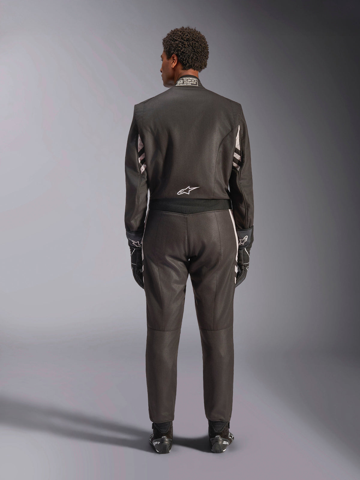 Alpinestars GP Race V3 Suit FIA, vista trasera, negro y plateado, mono de automovilismo profesional con construcción de aramida de dos capas y paneles elásticos, usado con guantes y botas de carreras a juego.