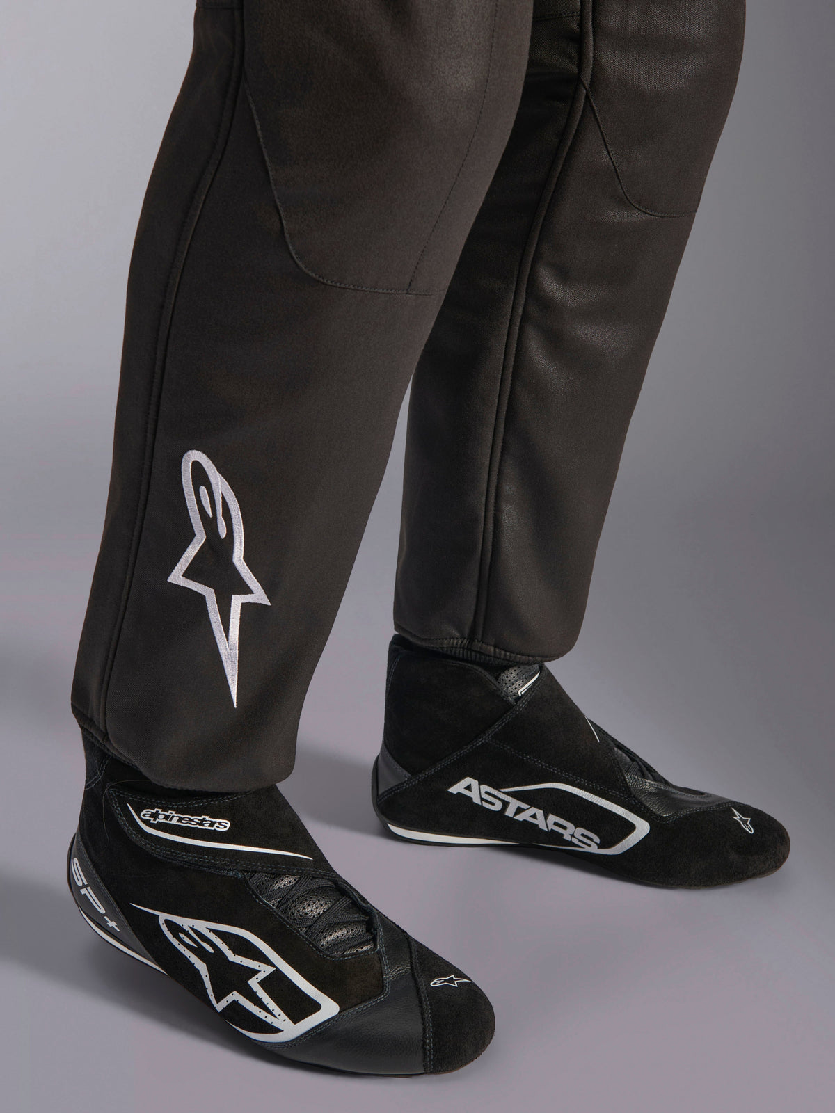 Alpinestars GP Race V3 Suit FIA, primer plano de la parte inferior de las piernas y botas de carreras, negro con detalles plateados, con el logotipo de la estrella bordado en blanco en la espinilla y puños acanalados, equipo de protección profesional para automovilismo.