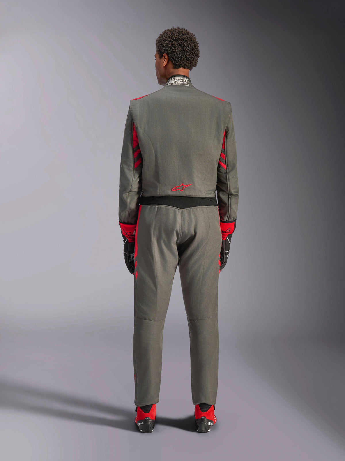 Vista trasera de un modelo vistiendo el mono Alpinestars GP Race V3 en Anthracite Red, traje de automovilismo profesional con detalles rojos en las mangas y las piernas, combinado con botas de carreras rojas y guantes negros, indumentaria técnica de automovilismo en un entorno de estudio.