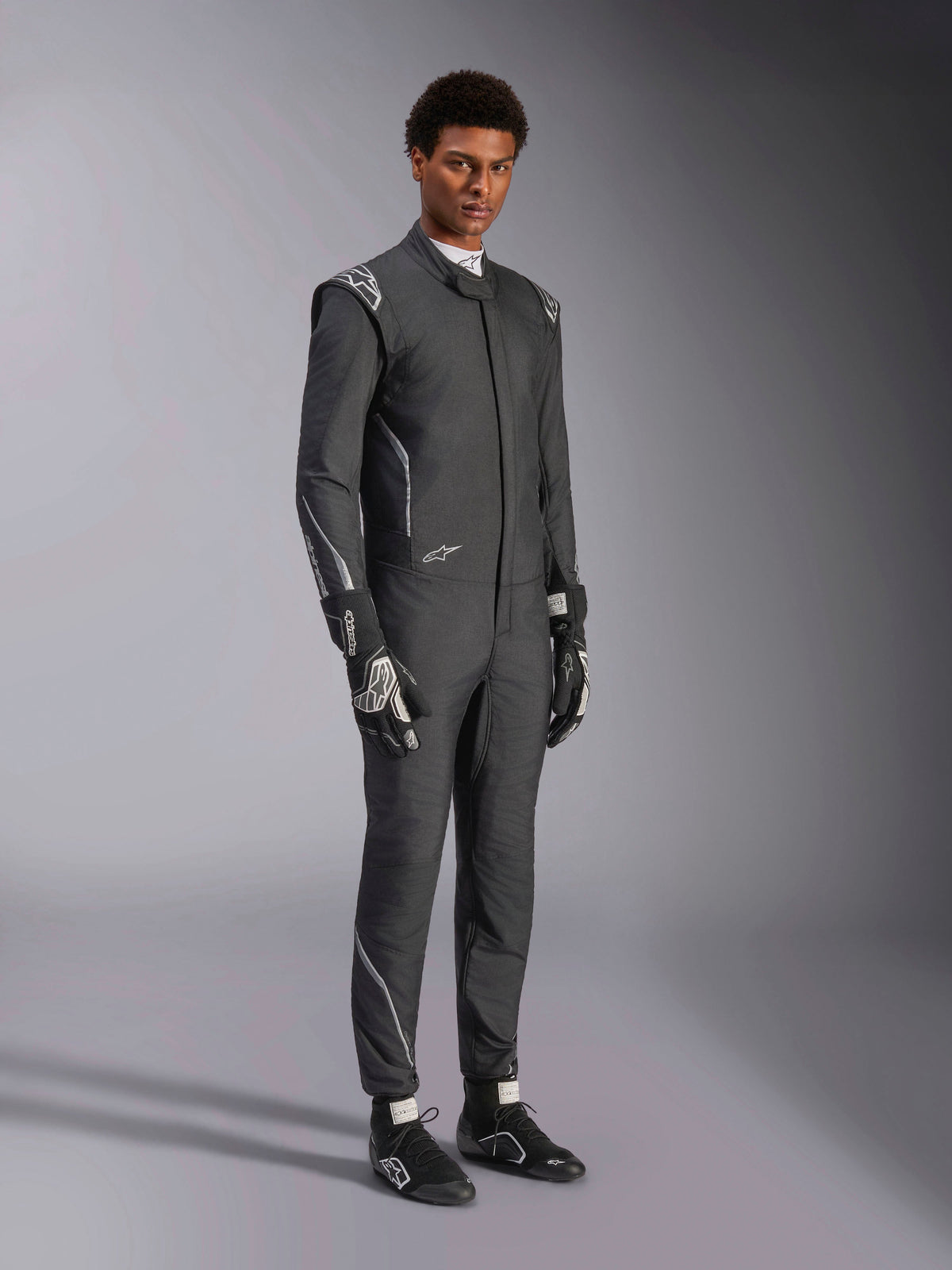 Mono Alpinestars Hypertech V3, traje de automovilismo, negro y Dark Shadow, usado por un modelo masculino con guantes y calzado de competición negros a juego, corte atlético ajustado, incluye logotipos de Alpinestars y detalles en gris claro, equipamiento de protección profesional para automovilismo.