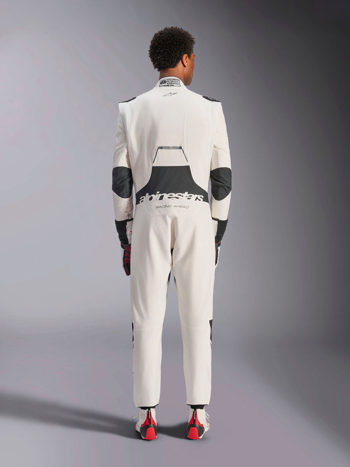 Vista trasera de un modelo luciendo el mono Alpinestars Hypertech V3 en blanco, negro y plata, mostrando el panel elástico lumbar negro con los logotipos 'alpinestars' y 'RACING AHEAD', inserciones negras en los codos, etiqueta de certificación FIA en el cuello y un corte atlético ajustado diseñado para la competición profesional.