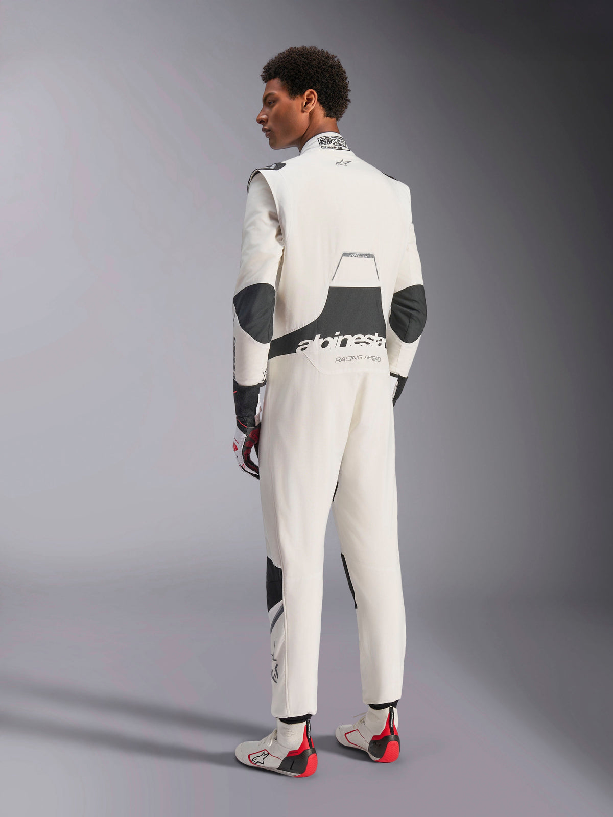 Mono Alpinestars Hypertech V3, traje de automovilismo profesional, blanco, negro y plateado, vista trasera del modelo mostrando paneles elásticos negros en la zona lumbar y los codos, logotipos de Alpinestars en la espalda, combinado con guantes y botas de carreras a juego para un look de automovilismo completo