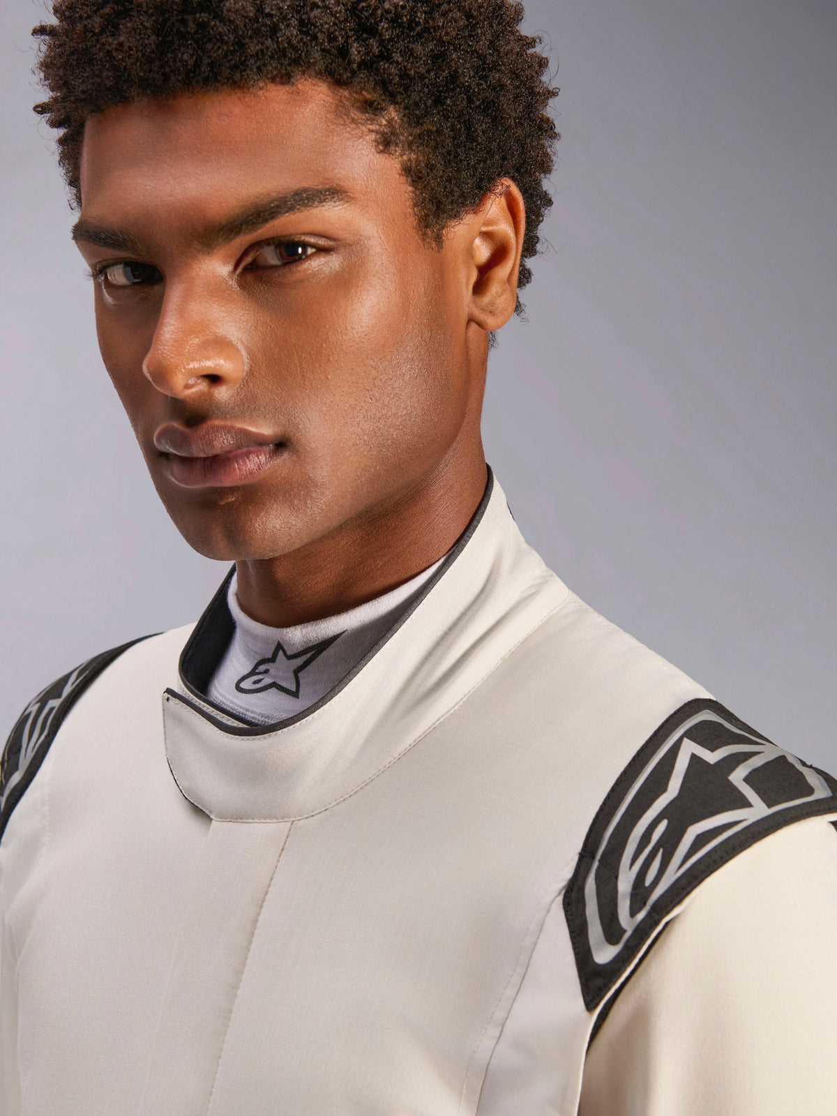 Primer plano de un modelo masculino con el mono Alpinestars Hypertech V3 en color blanco, negro y plateado, con un cuello alto con cierre seguro y logotipos de Alpinestars en los hombros, equipamiento profesional de automovilismo.