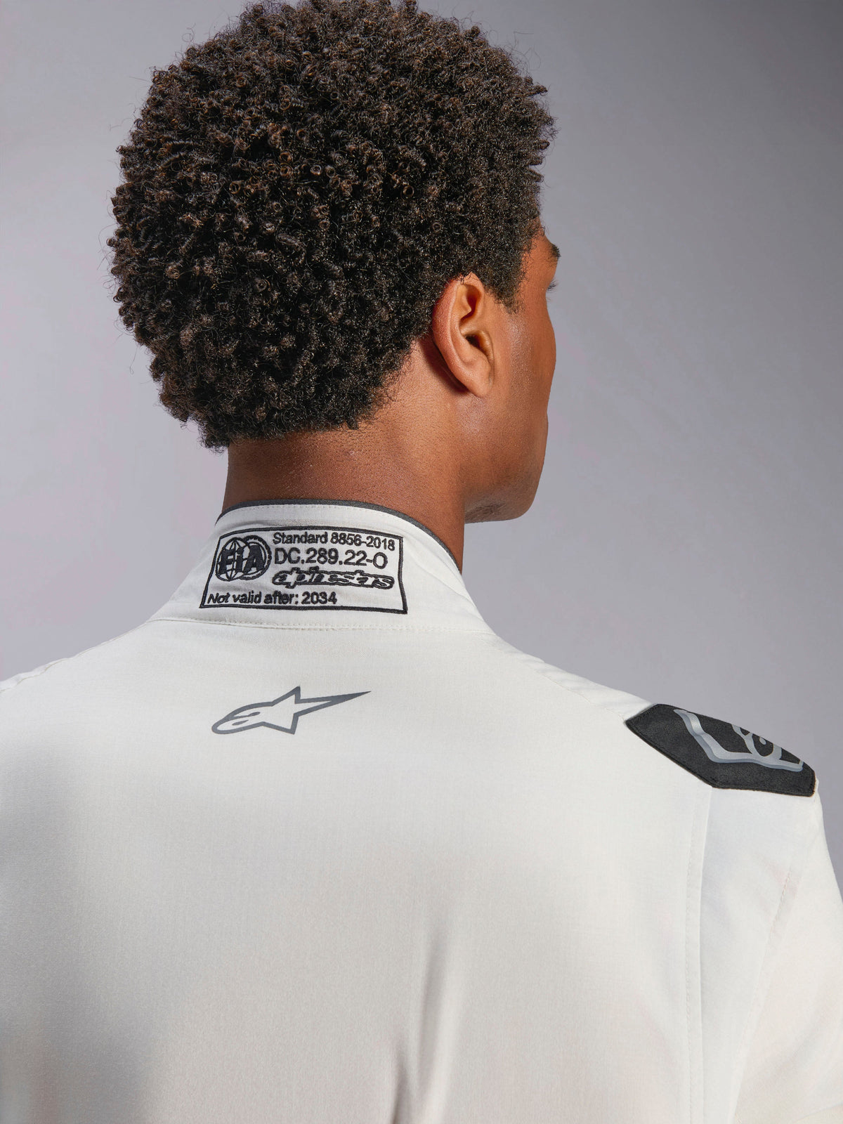 Mono Alpinestars Hypertech V3, blanco, vista de detalle del cuello posterior y el hombro, incluye etiqueta de certificación FIA 8856-2018, logotipo gris de Alpinestars impreso y asa de extracción negra, equipamiento de competición profesional