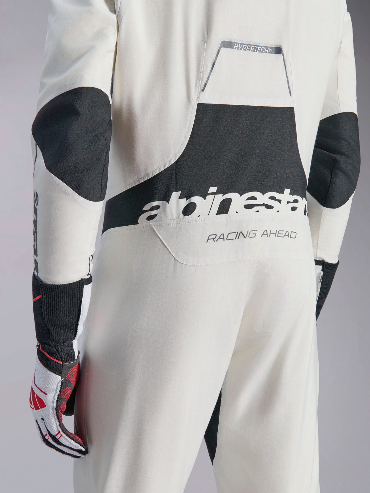 Alpinestars Hypertech V3 Suit, mono de automovilismo blanco, negro y plateado, vista posterior con gran logotipo negro de 'alpinestars' y texto 'RACING AHEAD', paneles elásticos en codos y zona lumbar, ropa de protección profesional para competición