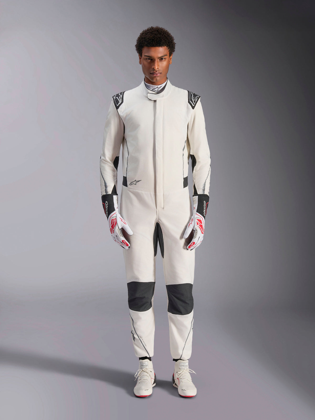 Mono Alpinestars Hypertech V3, mono de automovilismo profesional, blanco, negro y plateado, lucido por un modelo con guantes rojos y blancos y calzado de competición blanco a juego, con paneles de contraste negros y logotipos de Alpinestars, diseño de corte atlético ajustado