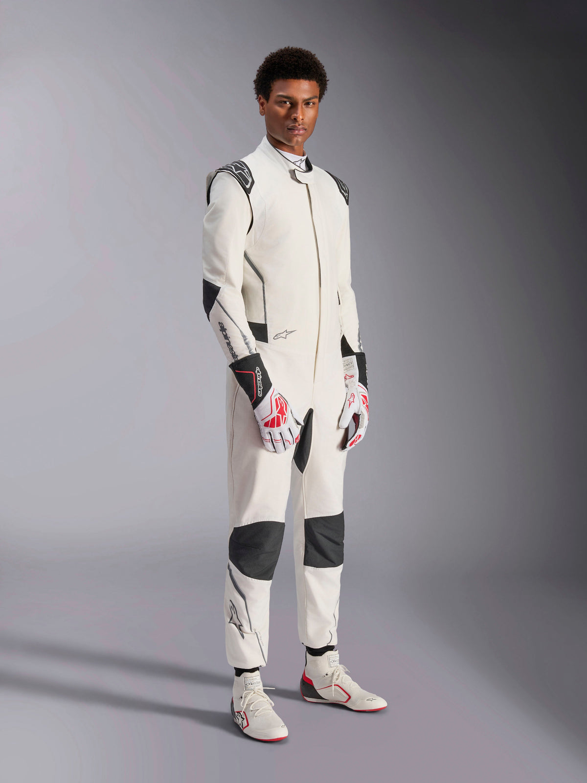 Modelo masculino con el mono Alpinestars Hypertech V3, blanco con detalles en negro y plateado, equipamiento profesional para automovilismo con paneles negros en los hombros y las rodillas, combinado con guantes de carreras blancos y rojos y botas de carreras blancas