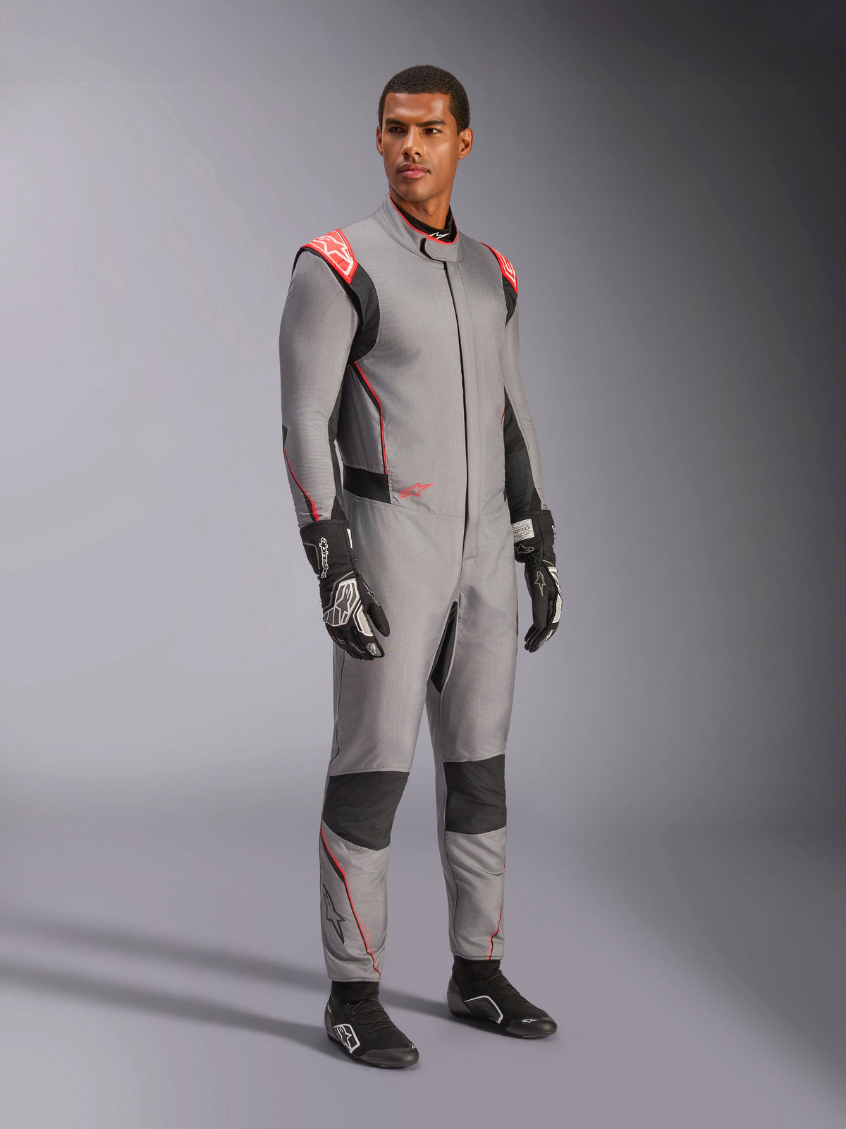 Modelo masculino con el mono Alpinestars Hypertech V3 en gris medio con acentos en negro y rojo flúor, traje de automovilismo profesional combinado con guantes y calzado de carreras negros, con confección ligera de dos capas de Nomex, detalles rojos en los hombros y un ajuste atlético