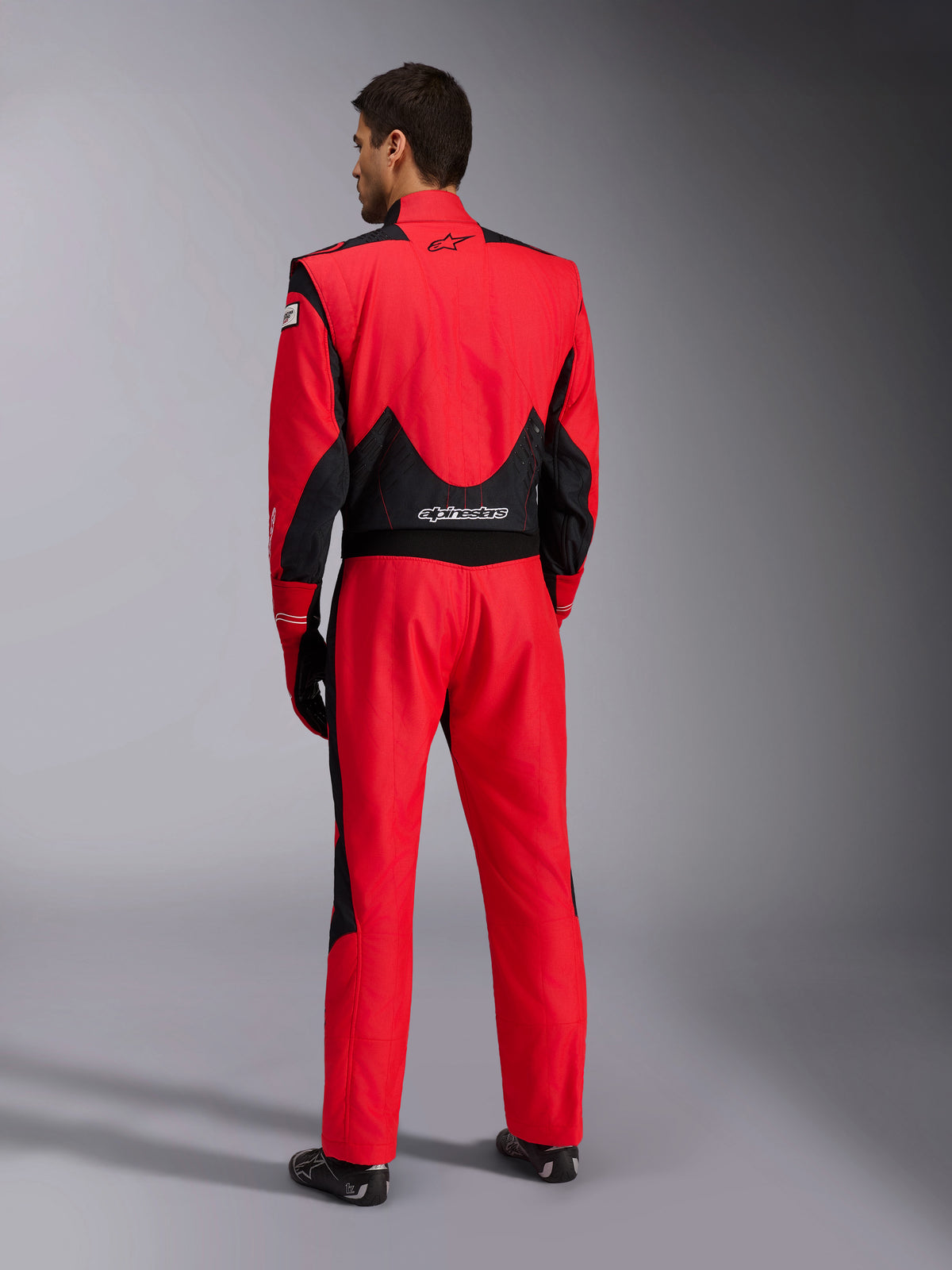 Vista posterior completa del traje de carreras Alpinestars GP Pro Comp V2 FIA, rojo y negro, con paneles de malla negra transpirable en los hombros y la parte inferior de la espalda, logotipo blanco de Alpinestars, usado con guantes y botas de competición a juego, equipamiento profesional para pilotos de automovilismo