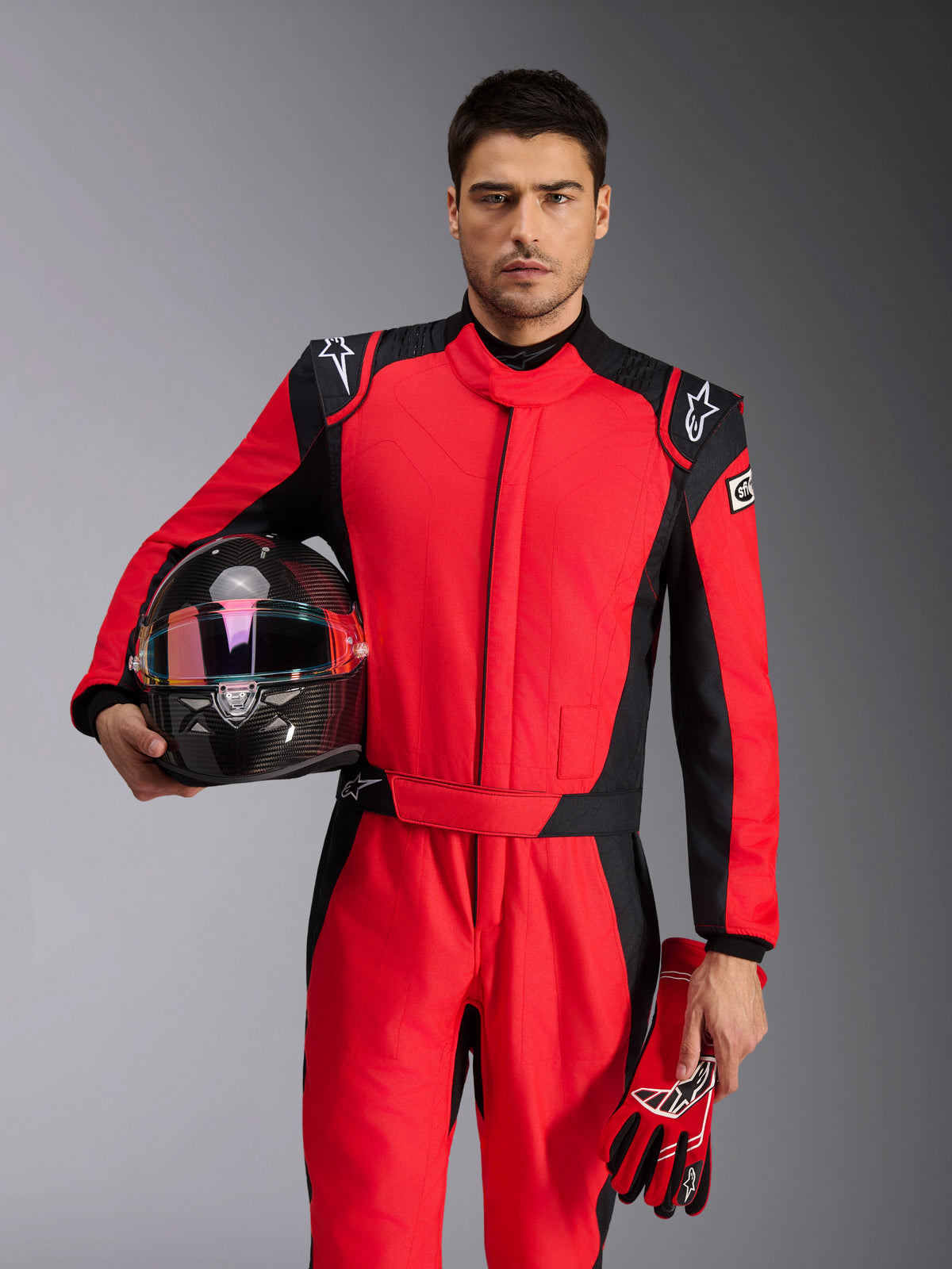 Piloto de carreras masculino con traje Alpinestars GP Pro Comp V2 FIA en rojo y negro, sujetando un casco de carreras de fibra de carbono y guantes a juego, vestimenta profesional de automovilismo