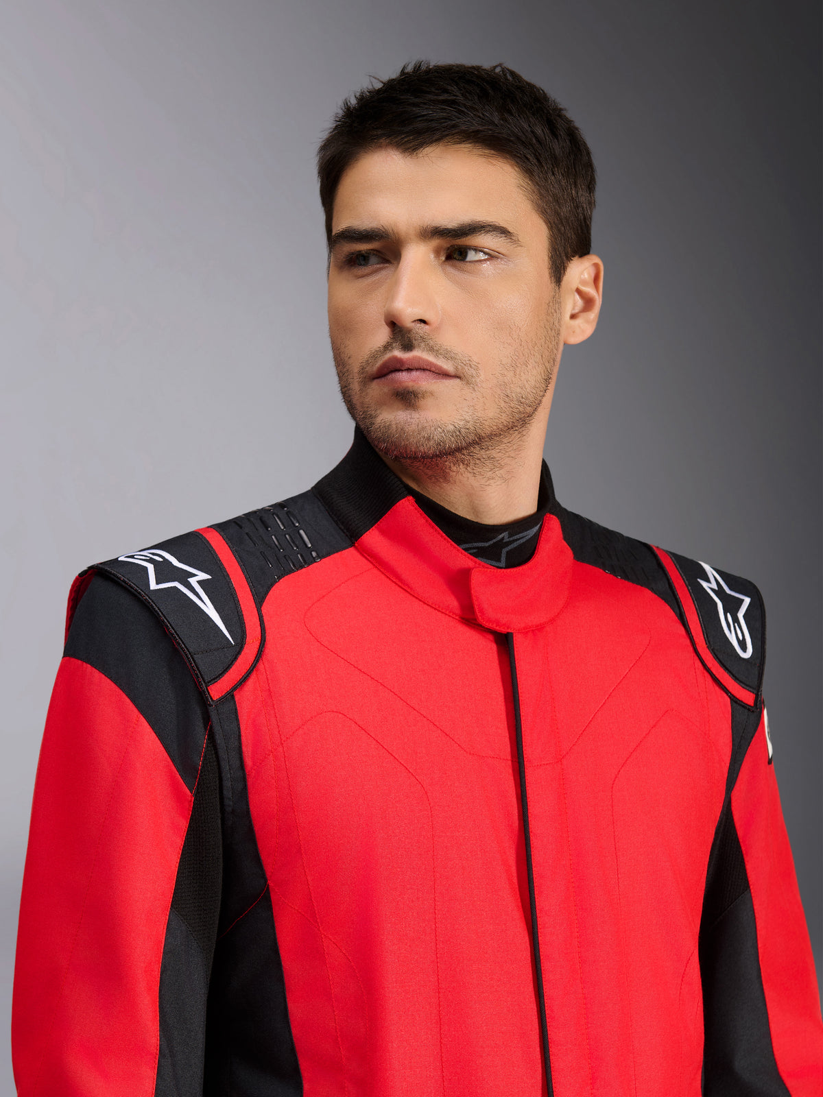 Modelo masculino vistiendo el traje de automovilismo Alpinestars GP Pro Comp V2 FIA en rojo y negro, con logotipos Astars blancos en los hombros y detalles de tecnología de agarre de silicona, indumentaria profesional de automovilismo