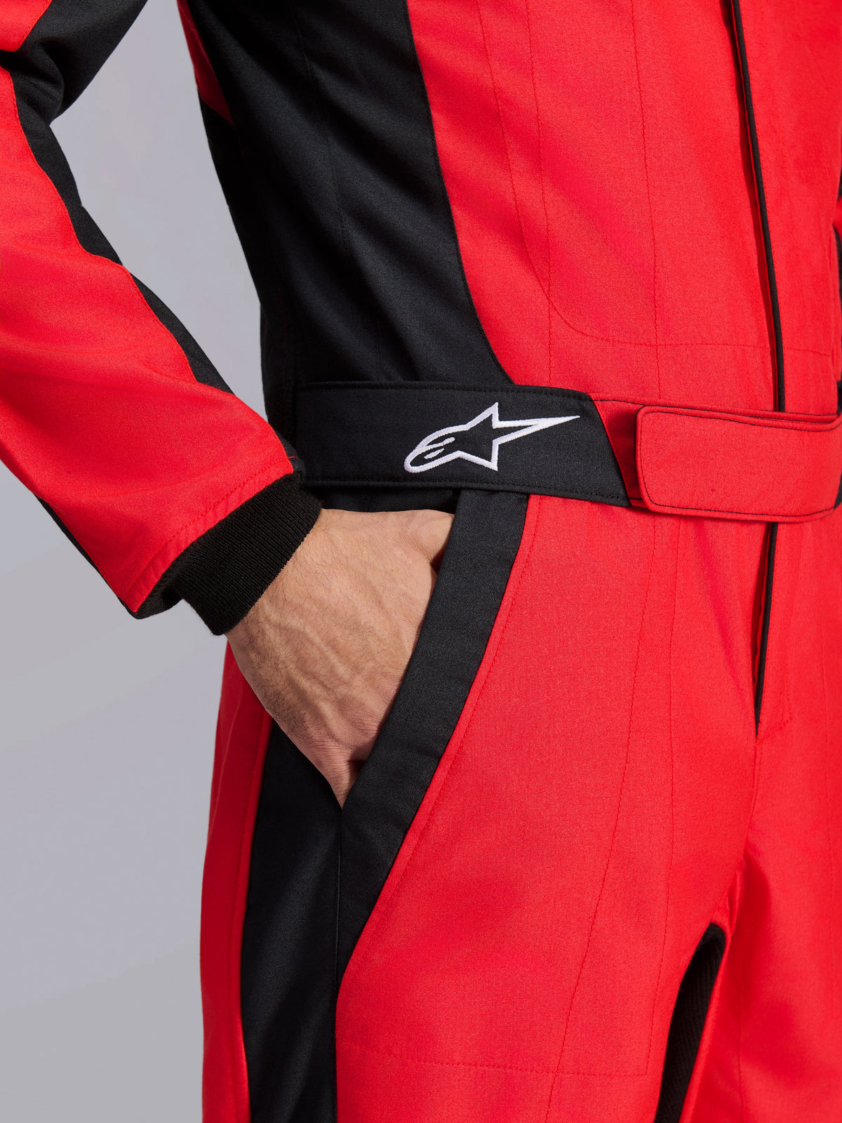 Traje de carreras Alpinestars GP Pro Comp V2 FIA, rojo y negro, primer plano de la zona de la cintura mostrando una mano en el bolsillo, con cinturón negro con logo de estrella blanca y paneles estructurados para automovilismo