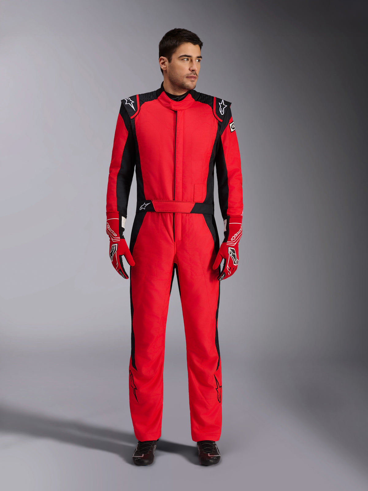 Modelo masculino luciendo el traje de automovilismo Alpinestars GP Pro Comp V2 FIA en rojo y negro, combinado con guantes de carreras rojos y calzado negro, mono profesional de una pieza con charreteras en los hombros y marca Alpinestars, equipamiento de alto rendimiento para pilotos