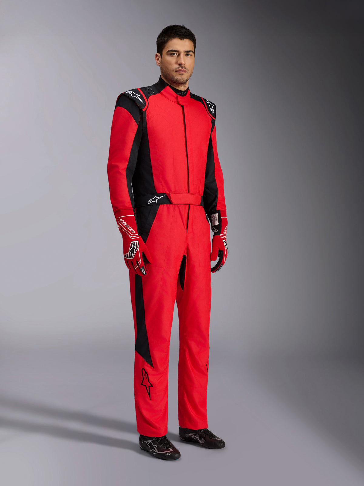 Traje Alpinestars GP Pro Comp V2 FIA, mono de automovilismo profesional en rojo y negro, usado con guantes rojos y blancos a juego y calzado de carreras negro, retrato de estudio de cuerpo completo de equipo de protección para deportes de motor.