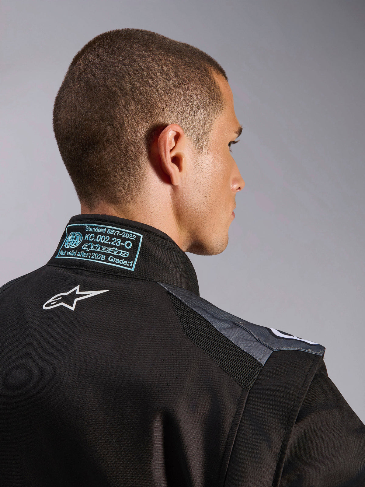 Alpinestars KMX-5 V3 Suit in Black and Dark Shadow, close-up view of the back and collar showing the white Astars logo, FIA 8877-2022 Grade 1 certification tag, and ventilated mesh shoulder panels.