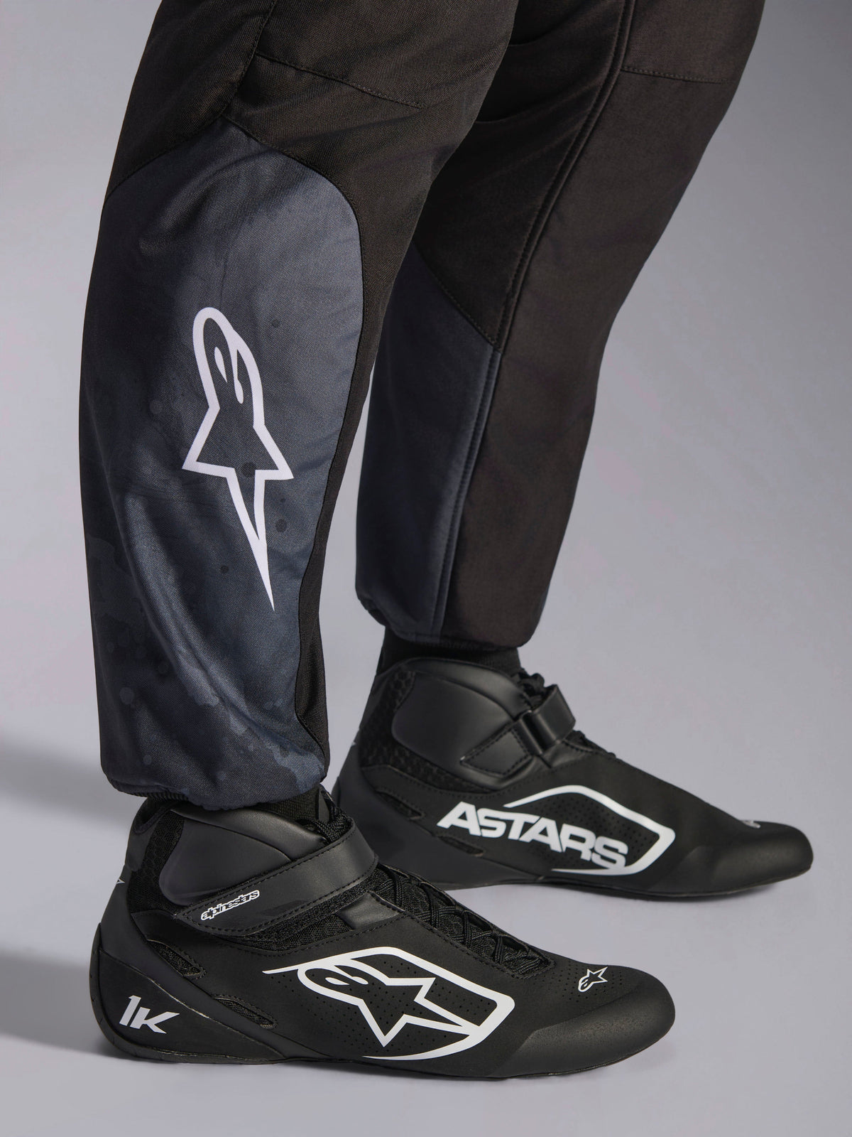 Alpinestars KMX-5 V3 Suit, Black Dark Shadow, close-up of lower legs and ankles, white star logo on calf, paired with black karting shoes, highlighting knitted ankle cuffs and athletic-regular fit.