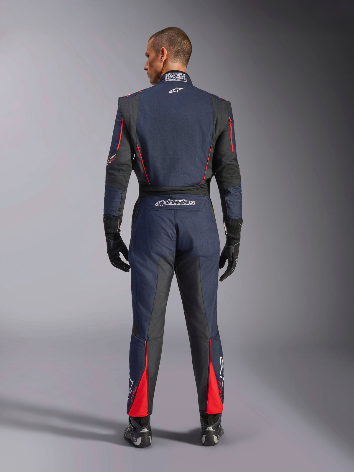 Rear view of the Alpinestars GP Tech V4 Suit in Blue Navy Black Red, one-piece auto racing suit with black stretch panels and red accents, featuring Alpinestars branding and FIA certification details, worn with professional racing gloves and boots.