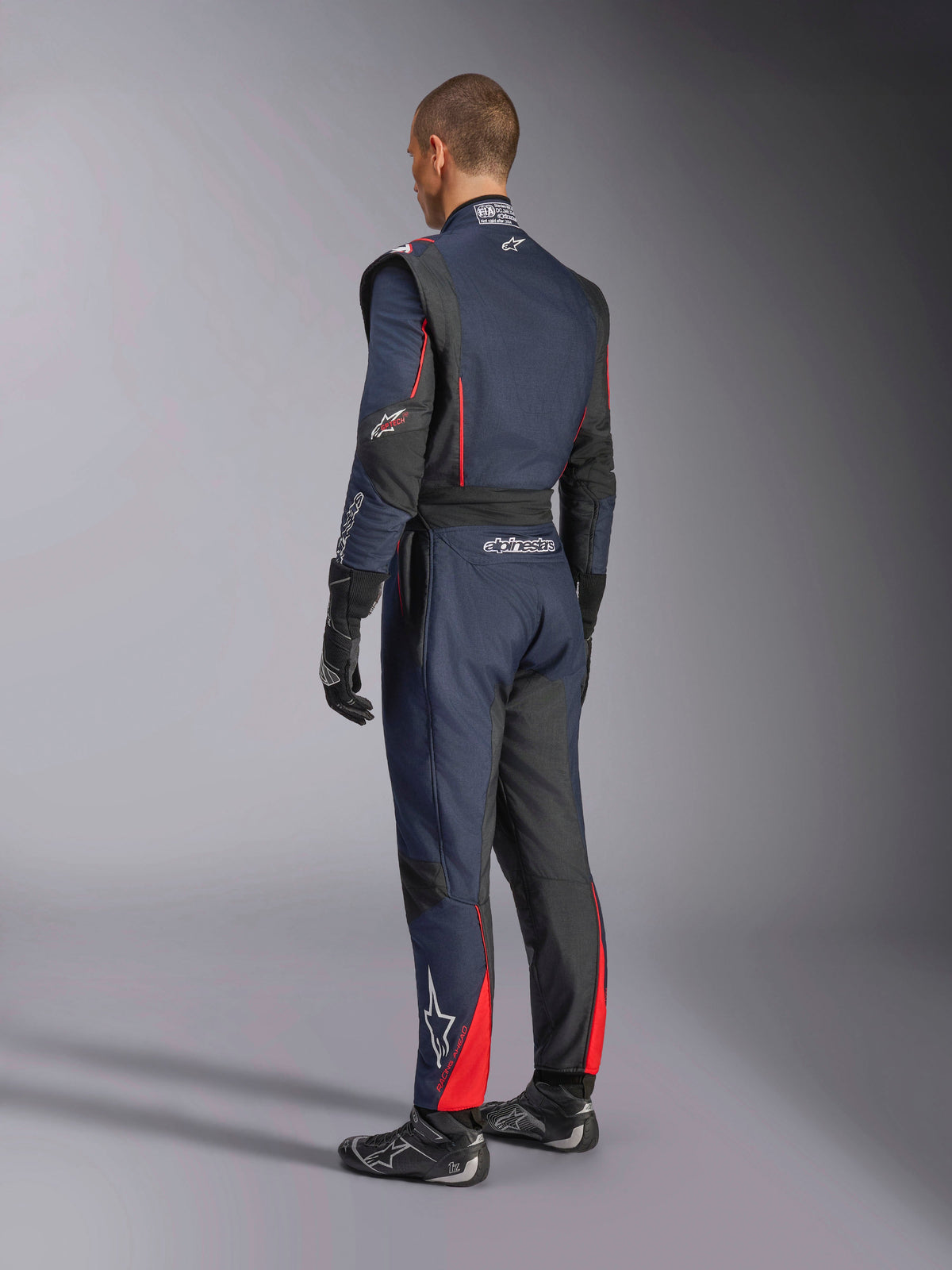 Alpinestars GP Tech V4 Suit, Blue Navy Black Red, back side view of professional one-piece auto racing suit, featuring red accents and black panels, worn with black racing gloves and shoes, FIA certified motorsport gear.