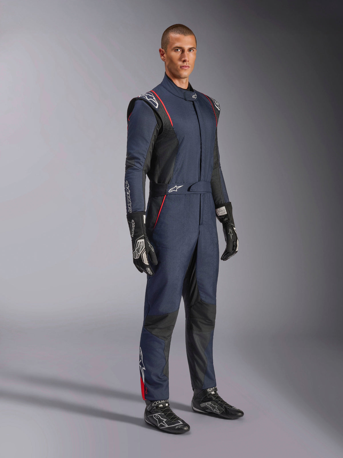 Alpinestars GP Tech V4 Suit, One-piece Auto Racing Suit, Blue Navy Black and Red, worn by a model with black racing gloves and boots, featuring black panels and red piping, professional motorsport gear.