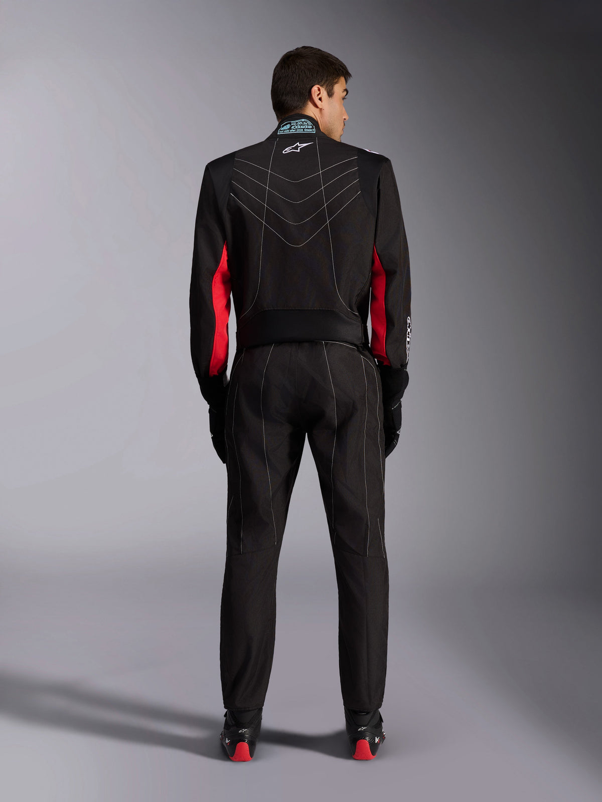 Mono de karting Alpinestars KMX-9 V3, negro con detalles en rojo y blanco, vista de cuerpo completo desde atrás, con costuras de contraste blancas en forma de V, paneles rojos en las mangas y logo de Alpinestars en el cuello, combinado con guantes y calzado de carreras negros