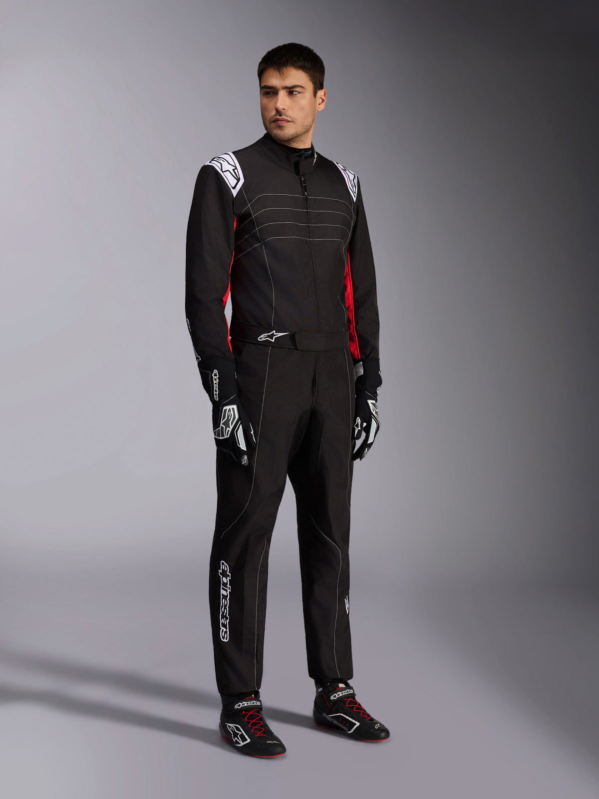 Mono Alpinestars KMX-9 V3, traje de karting negro, rojo y blanco lucido por un modelo, con hombreras y logotipos blancos, detalles laterales rojos, combinado con guantes y calzado de carreras a juego, ropa profesional para automovilismo