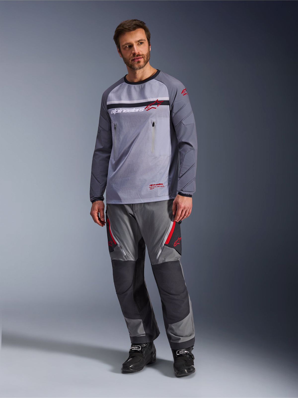 A man wearing the Alpinestars Maxdura Dual Jersey in Mid Gray and Dark Gray, styled with coordinating gray and black adventure pants and black boots, featuring a breathable mesh torso, zippered chest pockets, and red logo accents.