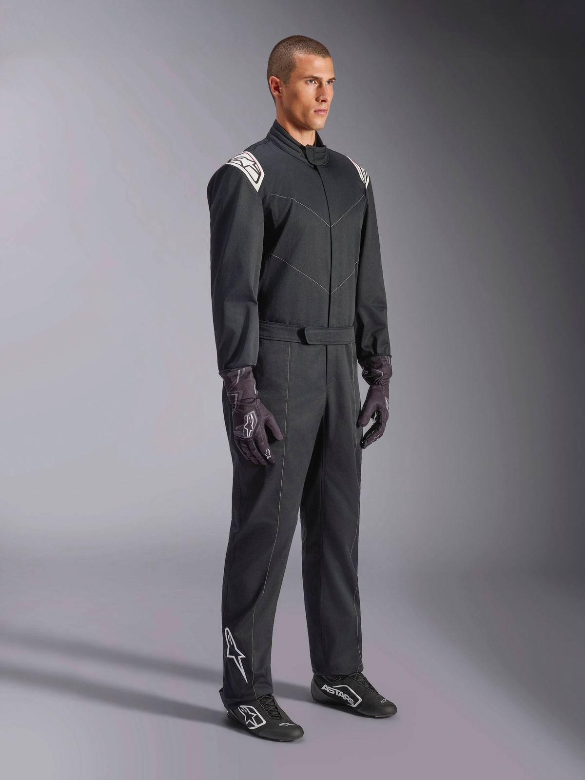 Alpinestars Kart Indoor V2 Suit, Black one-piece karting suit, worn by a model with racing gloves and shoes, featuring white shoulder accents and a white Astars logo on the lower leg, professional motorsport apparel for indoor track racing.
