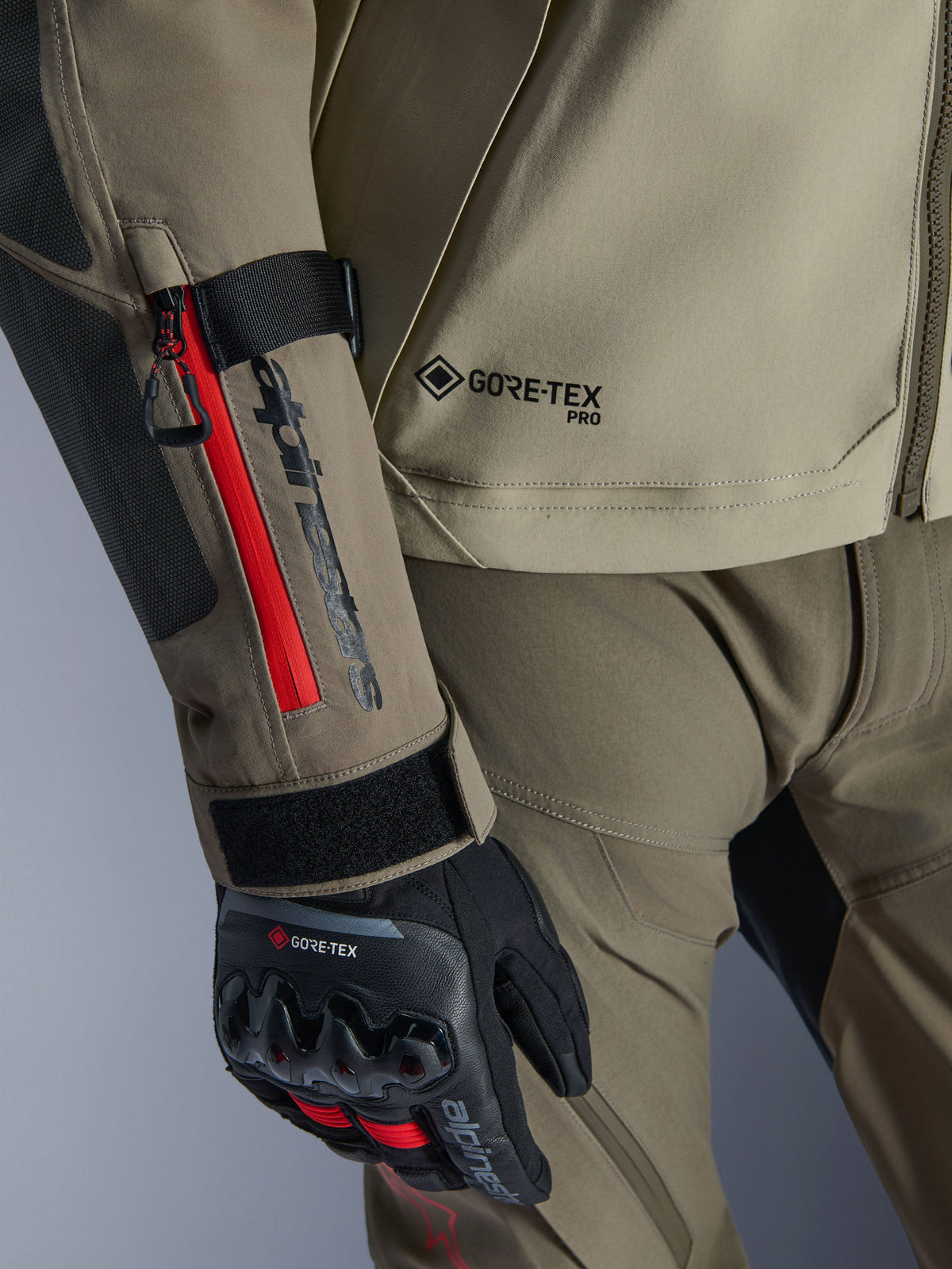 Close-up of Alpinestars Nazca 3L GORE-TEX PRO Jacket in Walnut Rock Brown, featuring red zipper accents on the sleeve, GORE-TEX PRO branding, and paired with black GORE-TEX adventure riding gloves.