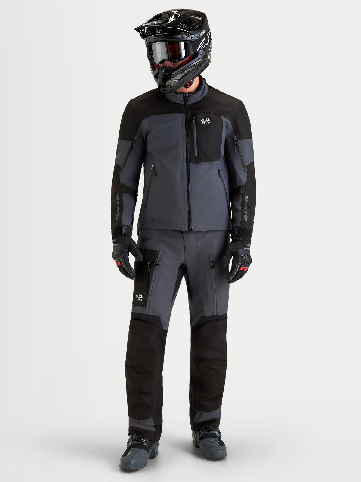 Alpinestars Arenis Gore-Tex Chaqueta, chaqueta de moto de aventura para hombre, en negro y gris oscuro, usada por un piloto con casco y equipamiento de protección a juego, vista frontal de cuerpo entero