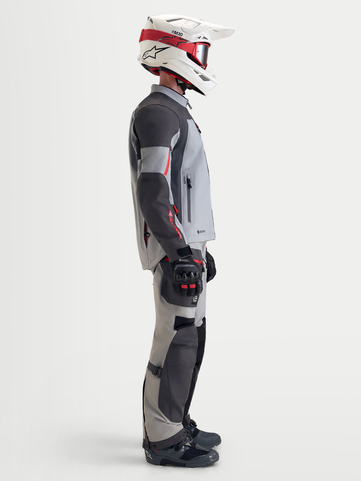 Alpinestars Arenis Gore-Tex Chaqueta, chaqueta de moto de aventura para hombre en gris medio, gris oscuro y negro, vista de perfil lateral de un piloto con la chaqueta textil impermeable, equipo a juego y casco blanco y rojo.