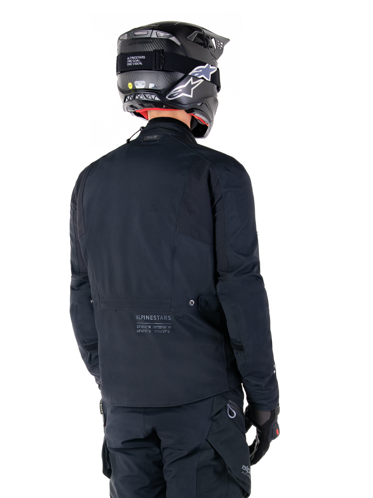 Chaqueta Alpinestars St-7 Prime GORE-TEX, negra, vista trasera de un piloto con la chaqueta, casco y pantalones a juego, con el logotipo de Alpinestars y coordenadas geográficas en la parte inferior de la espalda.