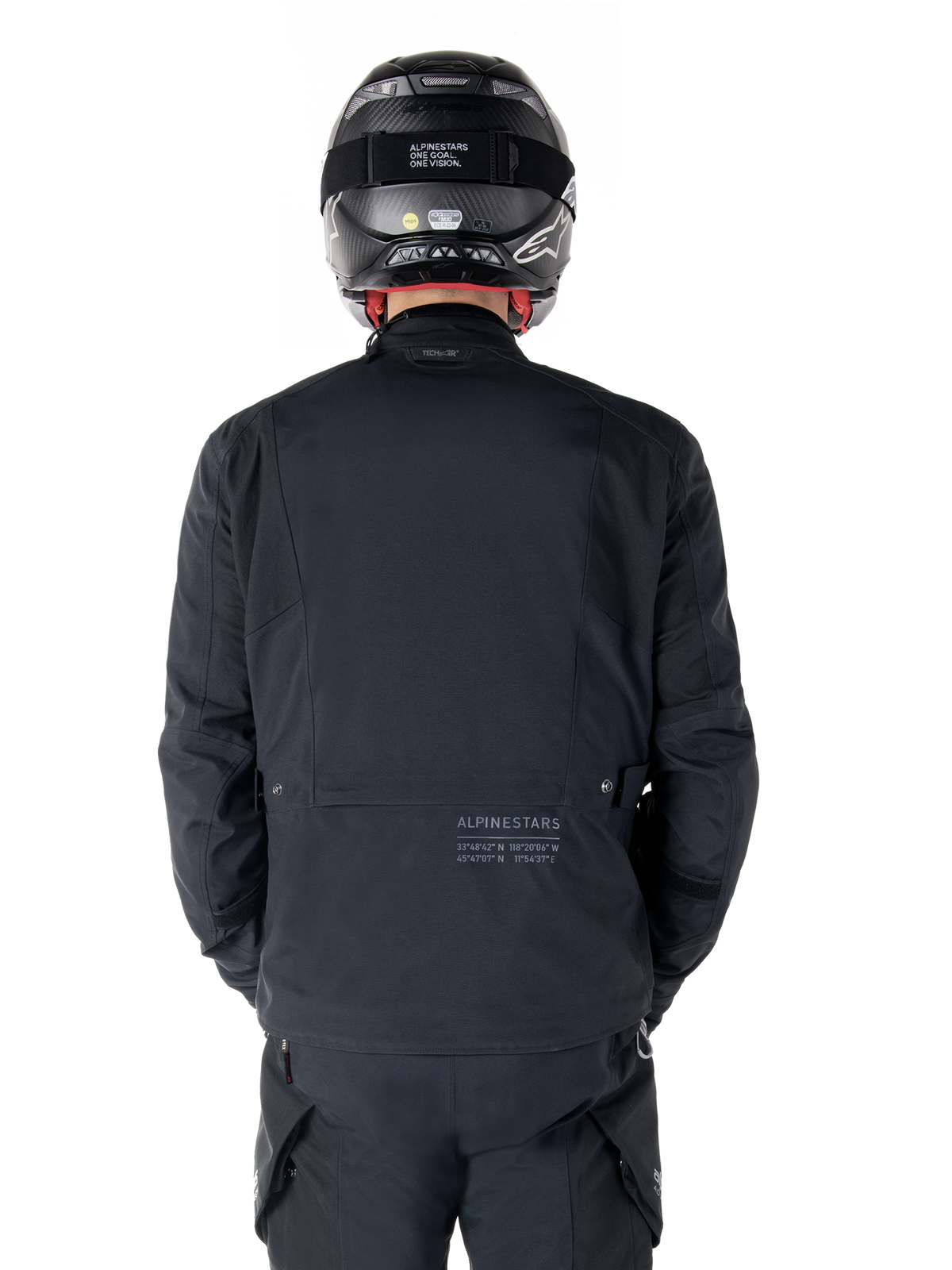 Chaqueta Alpinestars St-7 Prime GORE-TEX, negra, vista trasera de un piloto, con un logotipo sutil y coordenadas geográficas en la parte inferior de la espalda, combinada con casco y pantalones negros a juego, equipamiento de motociclismo elegante y técnico