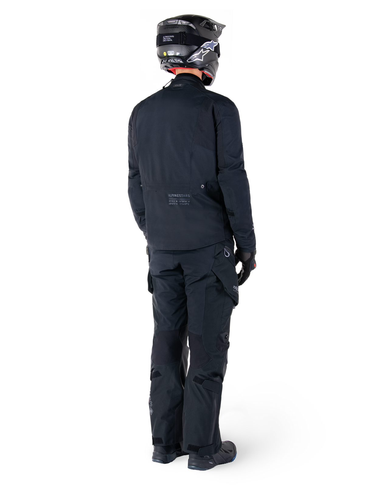 Vista posterior de un piloto con la chaqueta Alpinestars St-7 Prime GORE-TEX negra, combinada con pantalones de turismo negros a juego, casco de moto negro con logotipo blanco y botas negras, equipamiento de aventura completo para turismo y desplazamientos urbanos.