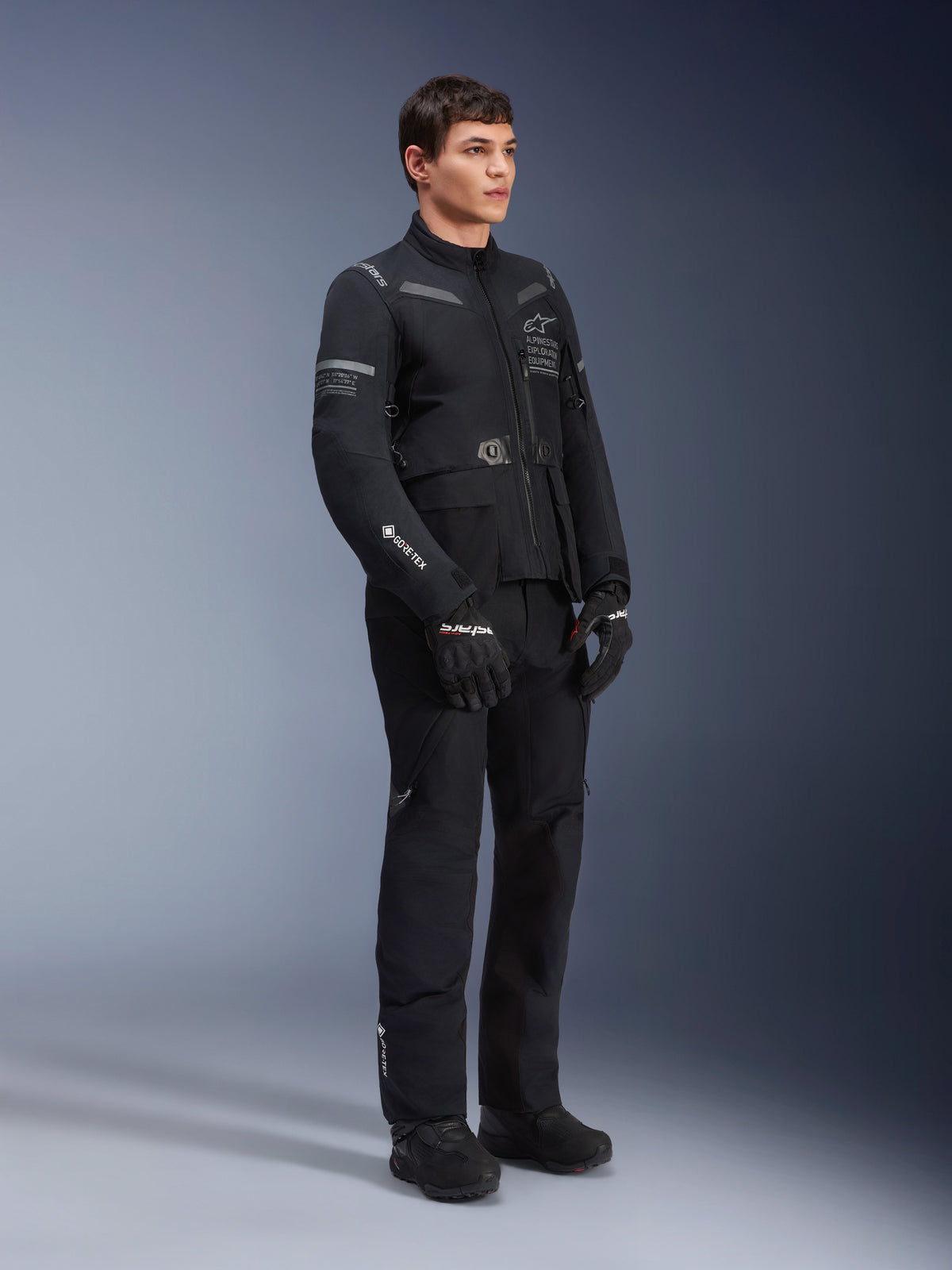 Pantalones Alpinestars ST-7 2L GORE-TEX, usados por un modelo con la chaqueta ST-7 a juego, conjunto de motociclismo adventure touring totalmente negro, con logotipos de GORE-TEX, bolsillos cargo, guantes y botas negros, foto de estudio sobre fondo gris