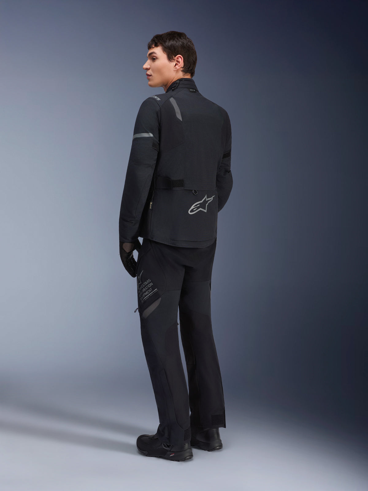Pantalones Alpinestars ST-7 2L GORE-TEX, pantalones de adventure touring, negro y gris oscuro, usados con la chaqueta ST-7 a juego y botas negras, con la marca 'Alpinestars Exploration Equipment' en el muslo, diseñados para una conducción impermeable y transpirable durante las cuatro estaciones.