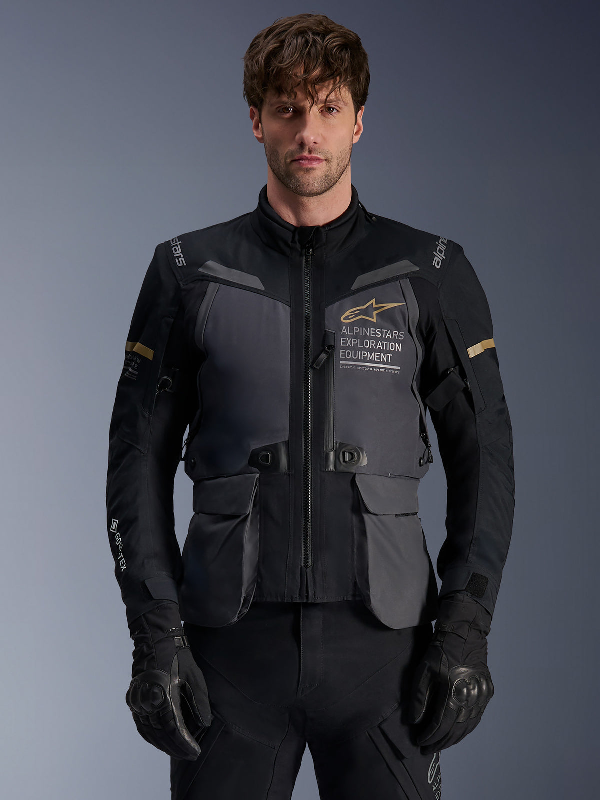 Chaqueta Alpinestars ST-7 2L GORE-TEX, chaqueta de moto Adventure Touring para hombre, gris oscuro y negro con detalles en caqui, con la marca 'Alpinestars Exploration Equipment', bolsillos cargo impermeables y cuello alto, usada por un modelo con guantes negros