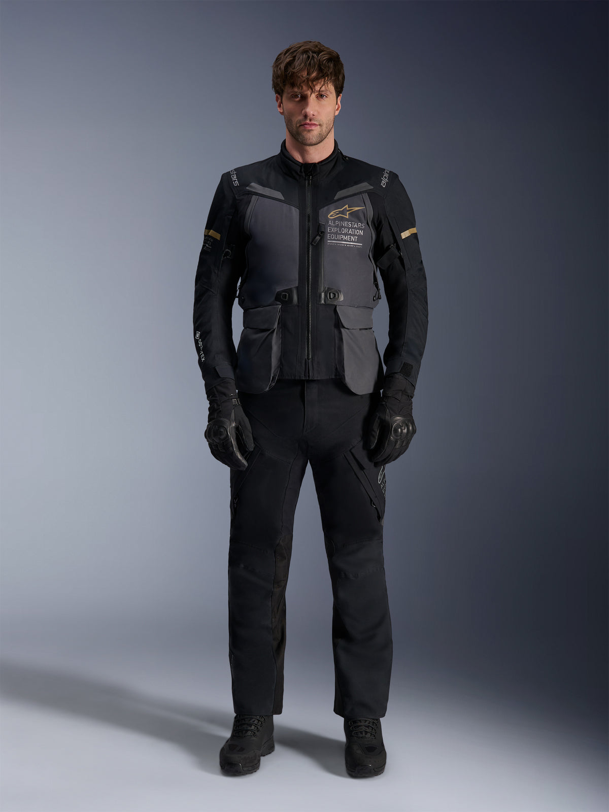 Modelo masculino con la chaqueta Alpinestars ST-7 2L GORE-TEX en gris profundo, negro y caqui oscuro, con el logotipo 'ALPINESTARS EXPLORATION EQUIPMENT' en el pecho, combinada con pantalones de pilotaje, guantes y botas negros a juego, vista frontal en estudio.