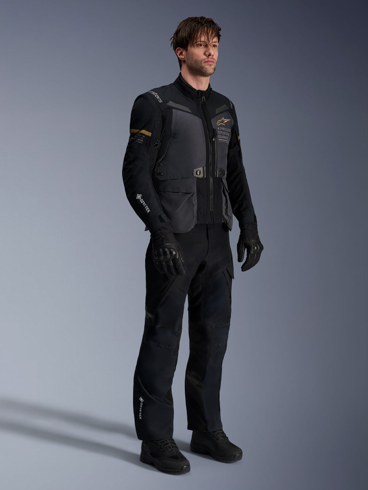 Modelo masculino con la chaqueta Alpinestars ST-7 2L GORE-TEX en gris oscuro negro y caqui oscuro, combinada con pantalones de aventura y guantes negros a juego, con capa laminada impermeable, bolsillos cargo y logotipos técnicos, ideal para adventure touring.