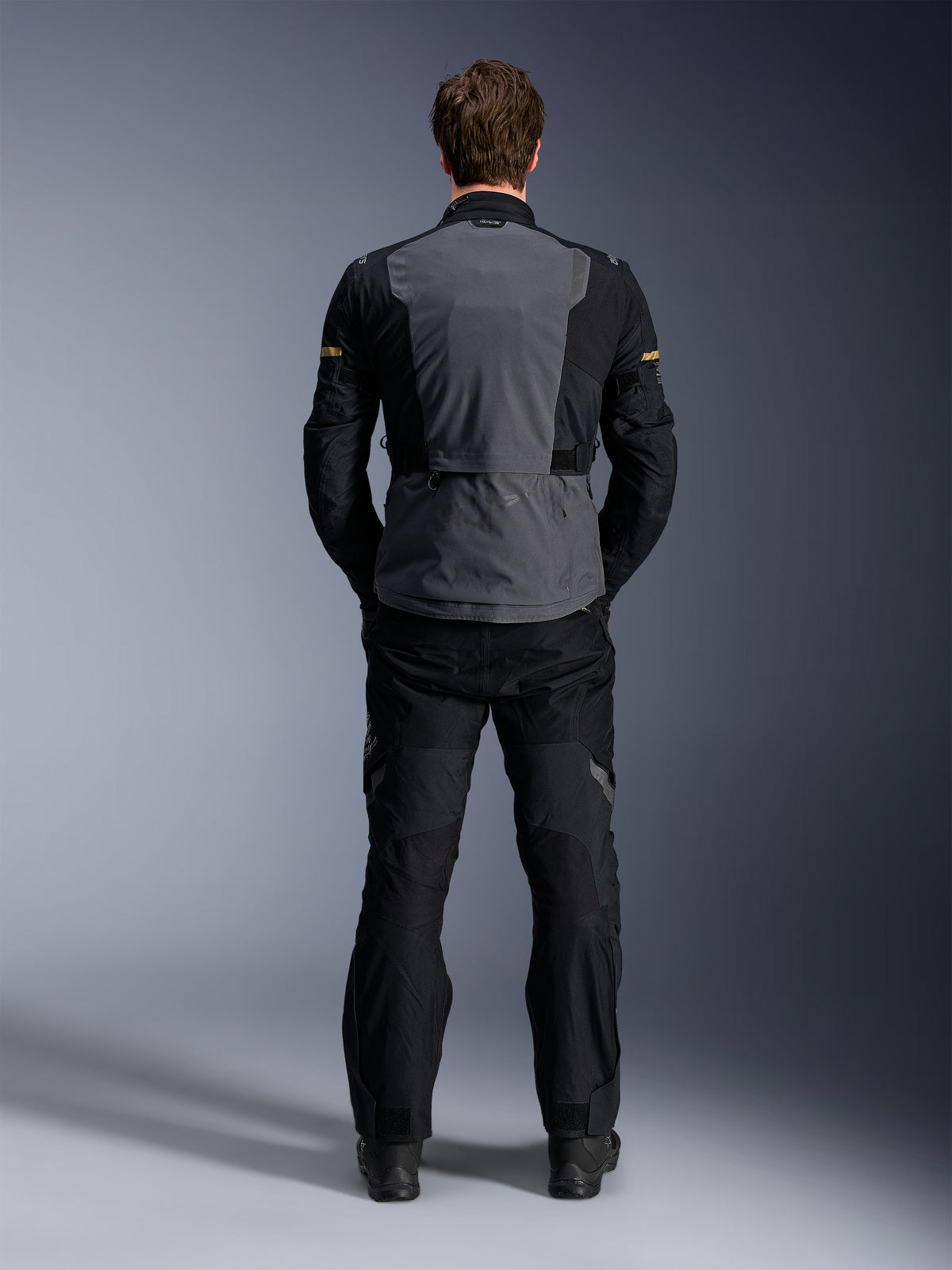 Chaqueta Alpinestars ST-7 2L GORE-TEX, Deep Gray Black y Dark Khaki, vista trasera en un modelo, chaqueta de moto para touring de aventura con panel trasero gris y mangas articuladas negras, combinada con pantalones negros a juego