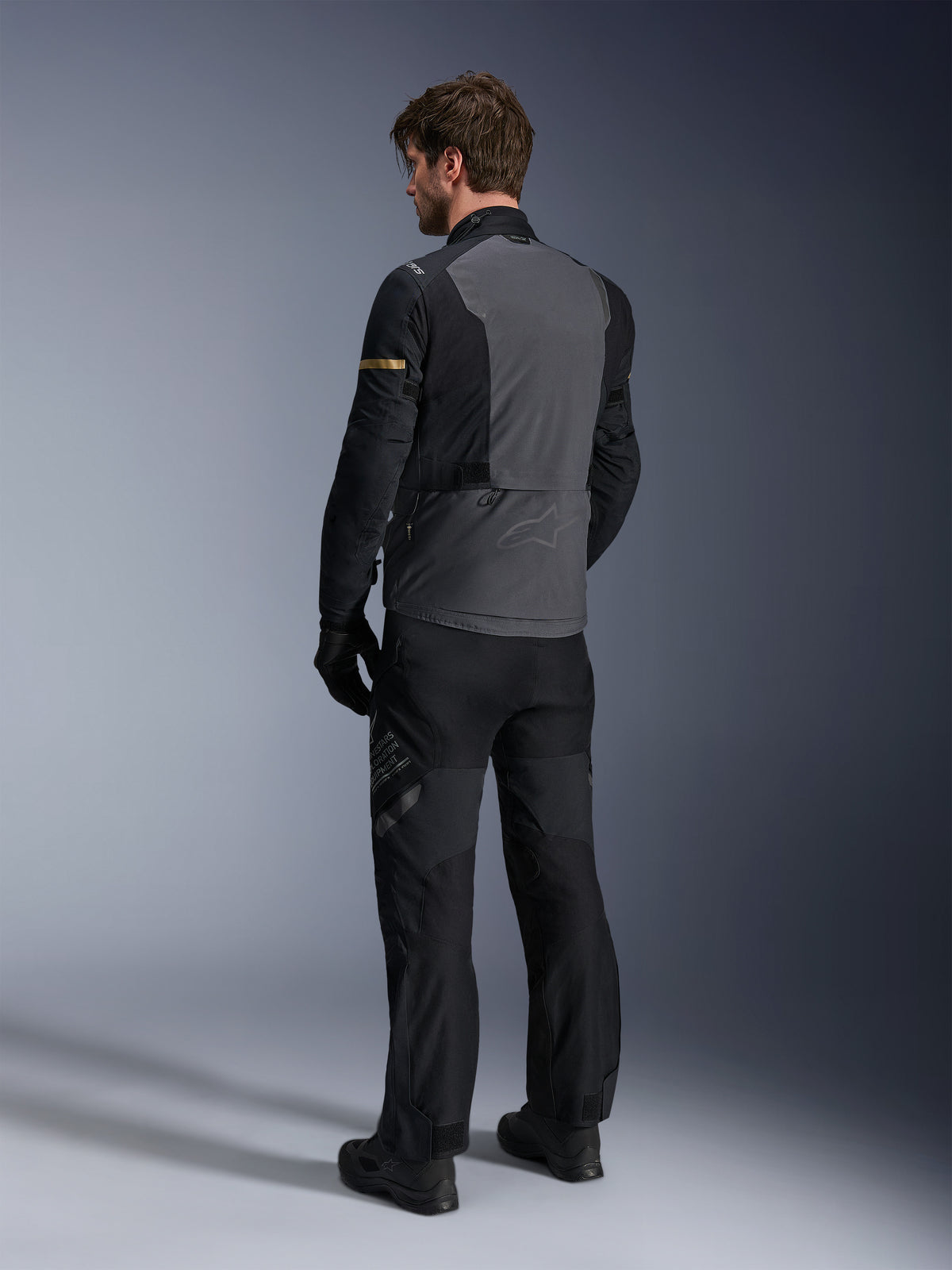 Chaqueta Alpinestars ST-7 2L GORE-TEX, gris oscuro y negro con detalles en caqui oscuro, vista posterior de un motociclista usando la chaqueta y pantalones de moto a juego, equipamiento de aventura touring con construcción laminada impermeable