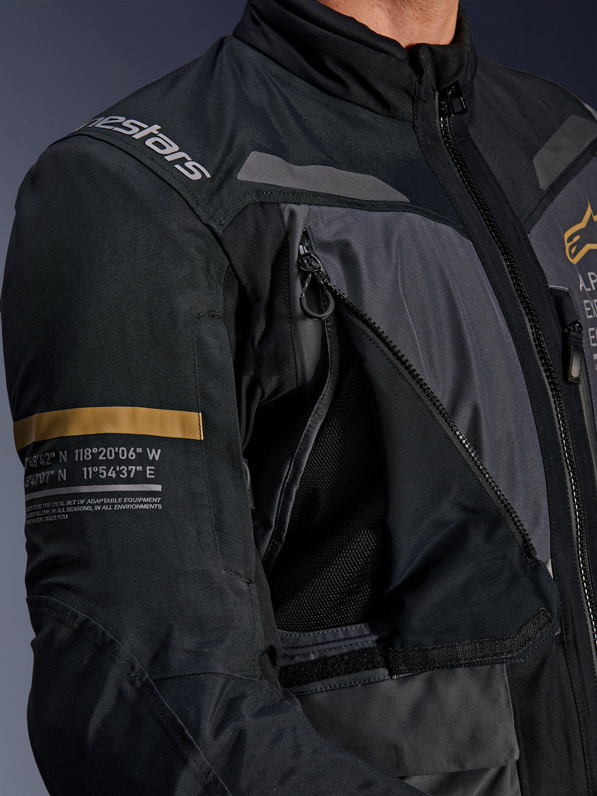 Chaqueta Alpinestars ST-7 2L GORE-TEX, gris oscuro y negro, primer plano que muestra el panel de ventilación del pecho abierto con cremallera y malla, logotipo de Alpinestars en el hombro y franja caqui en la manga con coordenadas geográficas impresas, equipamiento técnico de moto para adventure-touring