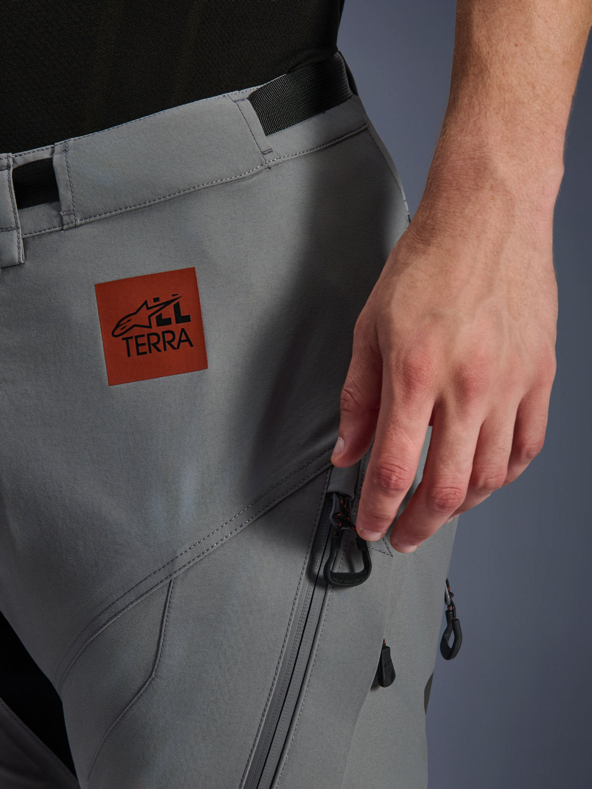 Pantalones Alpinestars Nazca 3L GORE-TEX PRO, pantalones de aventura para moto, color gris oscuro, primer plano de la parte superior del muslo con un parche del logo All Terra en marrón rojizo, bolsillo cargo con cremallera y tiradores negros, construcción textil duradera para conducción en todo terreno
